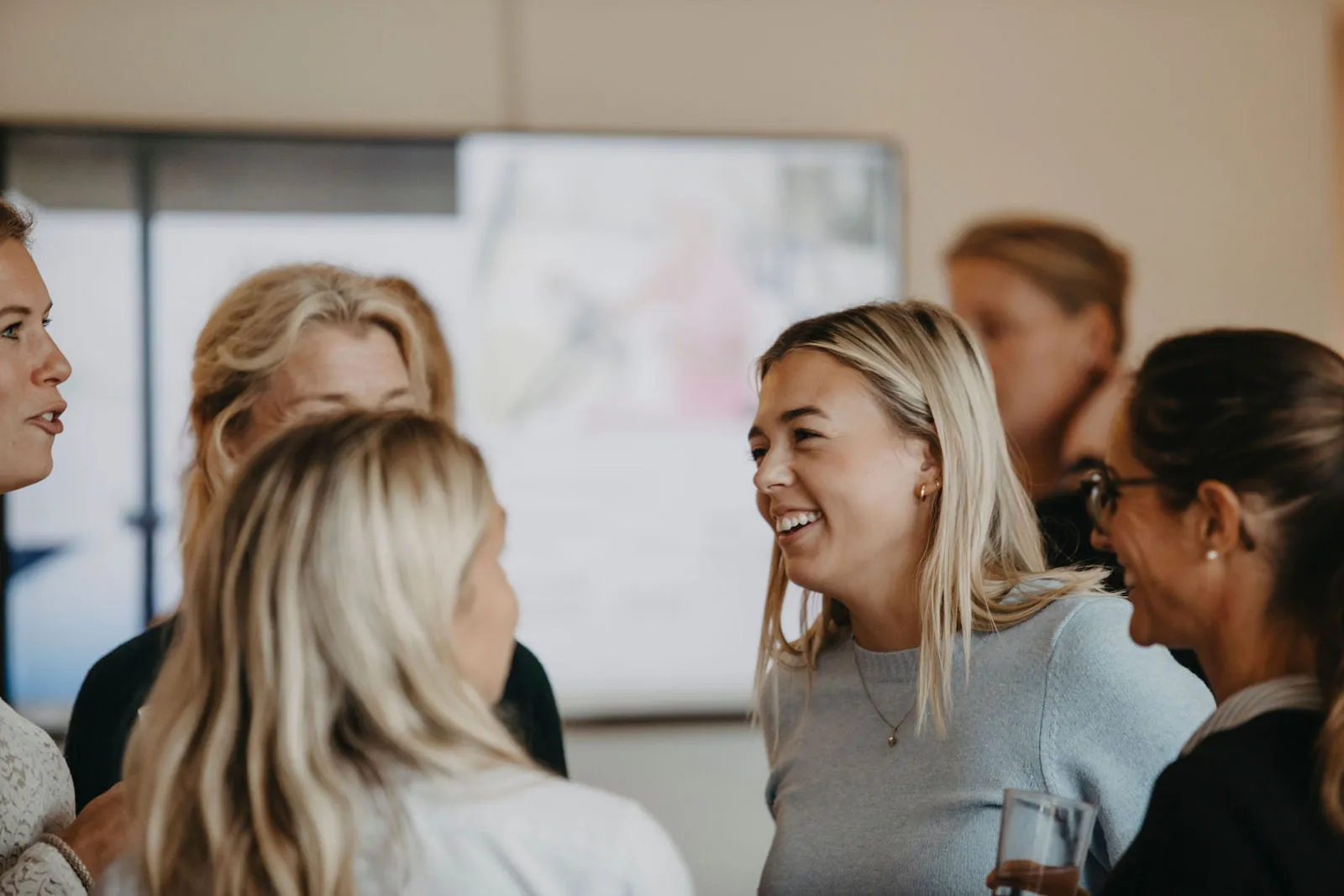 Fem glada kvinnliga medarbetare står och pratar med varandra i en grupp i ett konferensrum.